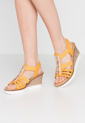 Gule kile-sandaler med flerfarvet flettet detalje på toppen og hvide teksturerede såler, båret på fødder mod en ensfarvet baggrund.