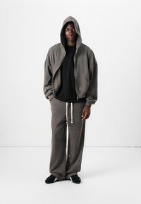 Gray zip-up hooded sweatshirt with large front pocket, paired with gray sweatpants featuring drawstring and decorative buttons. Black shirt underneath.