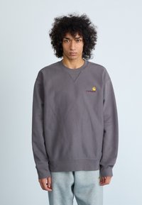 Sivi dukserica s rebrastim manžetama, koja na lijevoj strani prsnog dijela ima mali Carhartt logo u žutoj i crvenoj boji. Glatka tkanina, ležeran kroj.