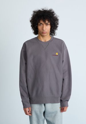 Sudadera gris con puños acanalados, que presenta un pequeño logo de Carhartt en amarillo y rojo en el lado izquierdo del pecho. Tejido suave, corte relajado.