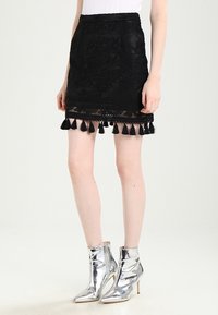 Černá vyšívaná mini sukně s krajkovým lemem a černými třásněmi podél spodního okraje; kombinováno se třpytivými stříbrnými kotníkovými botami.