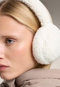 Vita fleeceöronmuffar med en mjuk, texturerad yta och ett plyschband, bärs bredvid ljust blont hår och en taupejacka.