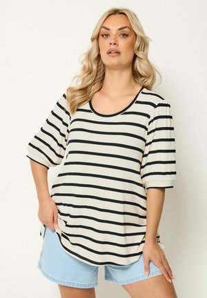 Femme blonde aux cheveux ondulés portant une chemise blanche à rayures horizontales noires et un short en denim bleu clair, debout devant un fond uni.