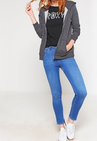 Felpa grigia con zip dal design leggero sopra una T-shirt grafica nera. Jeans blu aderenti e sneaker bianche completano l'outfit.
