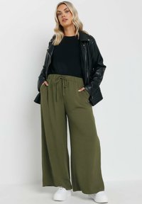 Olivgröna vida byxor med dragsko i midjan; kombinerat med en svart långärmad topp och en svart läderjacka. Vita sneakers.