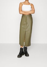 Jupe midi en cuir vert olive avec fente avant, associée à des bottes de combat noires à lacets. Haut crop blanc simple porté en dessous.