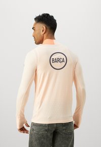Long-sleeved, light pink jersey with textured fabric and ribbed design; features a round navy "BARÇA" logo on the back.