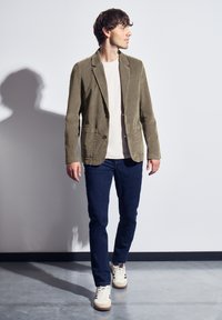 Americana de pana oliva con un solo botón y dos bolsillos frontales, usada sobre una camiseta ligera, combinada con jeans oscuros y zapatillas color crema.