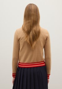 Cárdigan beige con un diseño ajustado, detalles de rayas rojas en los puños y el dobladillo, combinado con una falda azul marino con pliegues. Material de punto texturizado.