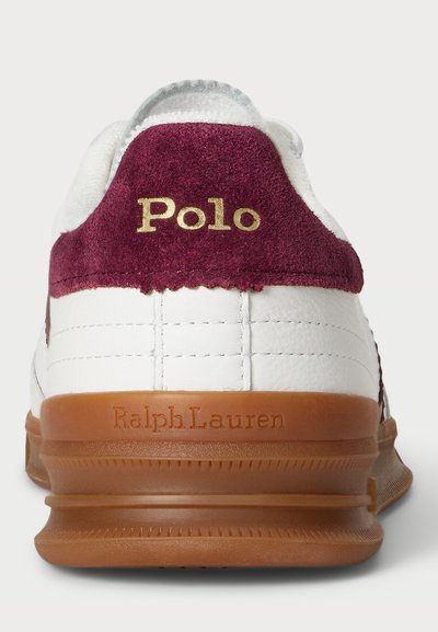 Witte sneaker met maroon suède hiel, gouden "Polo" logo en een bruine rubberen zool. Bevat geprofileerde stiksels op de bovenkant en merkdetails.