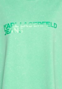 Světlá zelená látka s mírně vybledlým tučným textem "KARL LAGERFELD JEANS" v zelené barvě přes přední část.