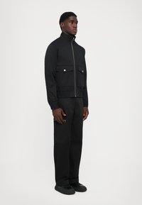 Jachetă neagră cu guler înalt, fermoar frontal, două buzunare pe piept cu capse și talie elasticizată. Pantaloni negri slim-fit completează aspectul.