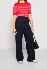 Punaisilla ja vaaleanpunaisilla raidoilla varustettu lyhythihainen neulepaita on yhdistetty korkeavyötäröisiin tummansinisiin leveälahkeisiin housuihin, ja asusteena on pieni musta käsilaukku.