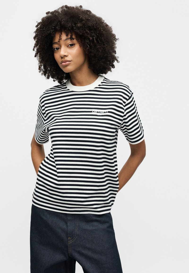 Kvinde med krøllet hår iført en sort og hvid stribet kortærmet skjorte og mørke jeans, stående med hænderne i lommerne mod en hvid baggrund.