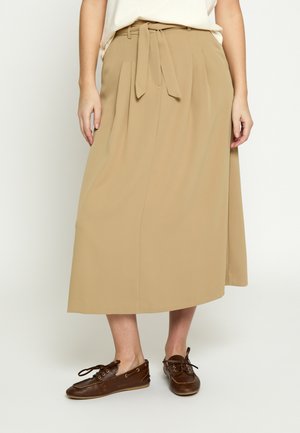 Woman wearing beige midi skirt with belt tied at waist and brown leather lace-up loafers, standing against a plain light background.