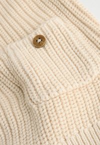 Pull en tricot crème avec texture côtelée et poche poitrine, fermée par un bouton rond marron. Fil épais et doux.