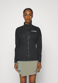 Schwarze Softshell-Jacke mit hohem Kragen, durchgehendem Reißverschluss und Terrex-Logo. Das Model kombiniert sie mit einem hellgrünen Rock, der Taschen hat.