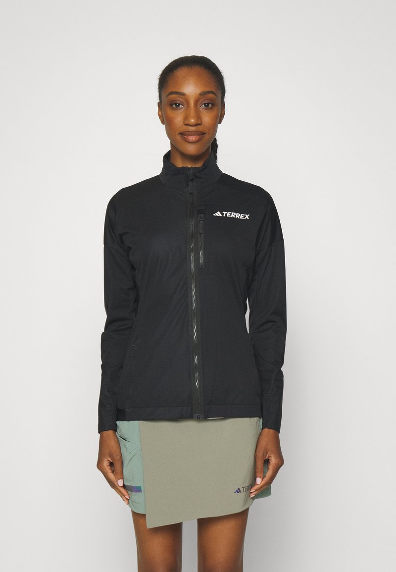 Schwarze Softshell-Jacke mit hohem Kragen, durchgehendem Reißverschluss und Terrex-Logo. Das Model kombiniert sie mit einem hellgrünen Rock, der Taschen hat.