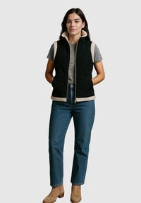Zwarte gewatteerde vest met beige accenten, hoge kraag, rits aan de voorkant, gecombineerd met een grijs T-shirt en blauwe jeans. Model staat met handen in de zakken.