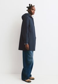 Marine vandtæt frakke med hætte, trykknapper og to sidelommer, parret med løstsiddende denimjeans og beige slip-on sko.