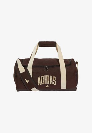 Brązowa torba sportowa Adidas z kremowymi uchwytami i paskiem na ramię, suwakiem z przodu oraz kremowym logo Adidas z boku.