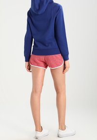 Felpa con cappuccio blu e tasca a marsupio, abbinata a pantaloni corti rossi con un motivo testurizzato e profili bianchi, indossata con sneakers bianche.