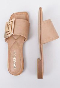 Sandalias deslizables en nude con una tira ancha y un acento geométrico dorado. Material suave, tacón bajo y plano, y detalles de costura visibles.