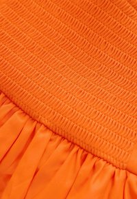 Oranje jurk met een gesmokt lijfje en een geplooide rok. De stof is soepel en heeft een gestructureerd horizontaal patroon op het bovenste gedeelte.