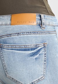 Jeans de mezclilla azul claro con una etiqueta de cuero marrón que dice "Noisy May" en la parte trasera de la cinturilla. Presenta acentos cosidos y un aspecto ligeramente desgastado.