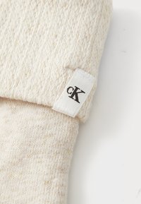 Közelkép egy krémszínű kötött anyagról, rajta egy fehér címkével, amelyen fekete "cK" logó található, a szélénél varrva.
