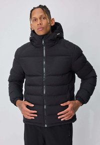 Veste matelassée noire avec une fermeture éclair à l'avant et une capuche. Comprend des sections matelassées, des manches longues et des poignets élastiques. Tissu lisse et résistant à l'eau.