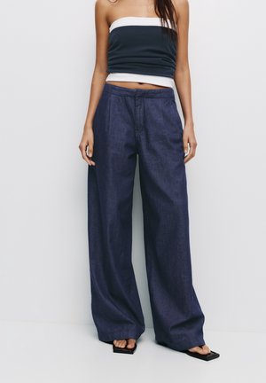 Femme portant un haut bustier bleu marine et blanc, un jean bleu foncé à jambes larges et des sandales noires ouvertes, debout devant un fond uni.