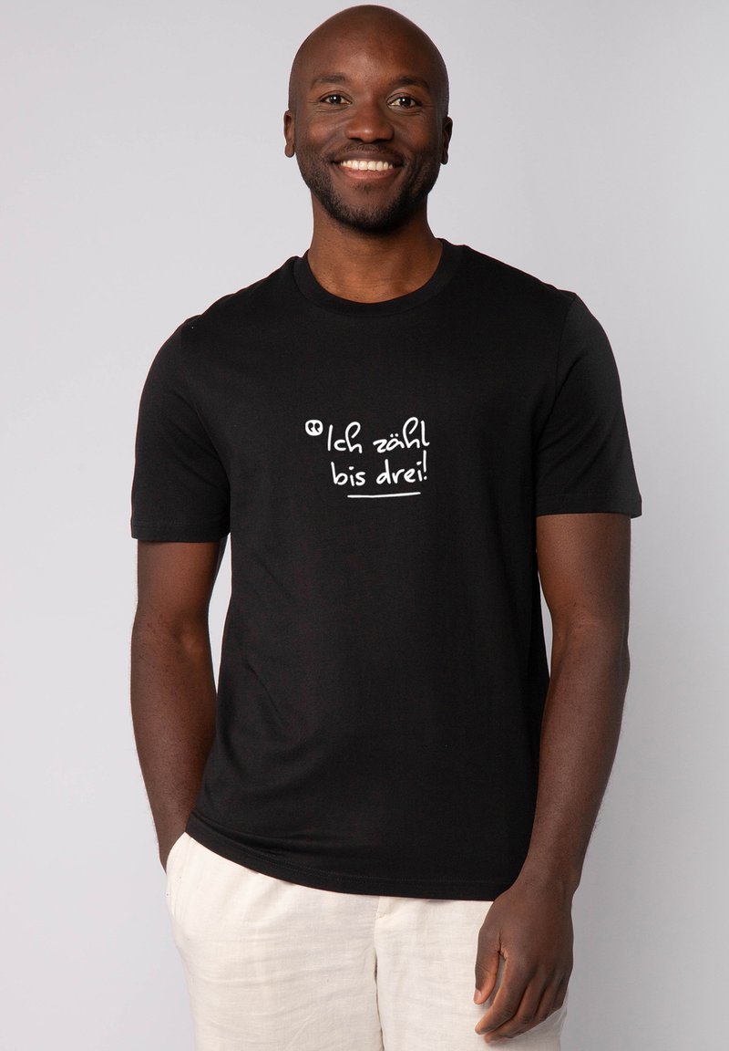 Homme souriant portant un t-shirt noir avec un texte blanc en allemand qui dit "Ich zähl bis drei !" et un pantalon clair.