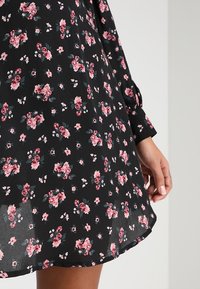 Černé šaty s květinovým vzorem, zdobené růžovými a zelenými květy, s dlouhými rukávy a volným střihem. Lehké, průsvitné tkaniny.