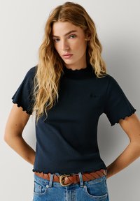 Μπλε ναυτικού χρώματος μπλουζάκι με ριπ υφή, διακοσμημένο με κυματιστά μανίκια και τελείωμα, με μικρό λογότυπο. Συνδυασμένο με καφέ πλεκτή ζώνη και ανοιχτό μπλε τζιν παντελόνι.