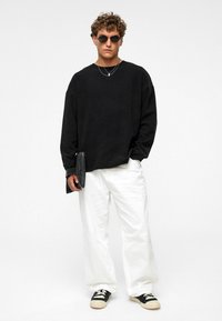 Zwarte oversized sweatshirt, witte wijde broek, zwarte zonnebril, gelaagde zilveren kettingen, houdt een zwarte clutch vast. Gelaagd stof, relaxte pasvorm.