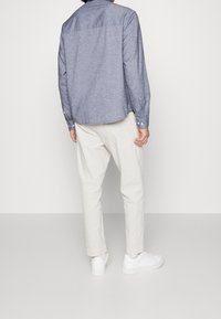 Modrá chambray košile s dlouhým rukávem a zaobleným lemem, s knoflíkovými manžetami, nošena s světle béžovými kalhotami a bílými teniskami. Zadní pohled.