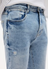 Svjetloplave traperice s lagano izblijedjelim izgledom, s istaknutim zakrpama i vidljivim šavovima. Džepovi s obje strane.