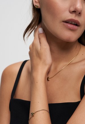 Woman wearing black top with matching gold heart-shaped earrings, necklace, and bracelet, gently touching her neck with her hand.