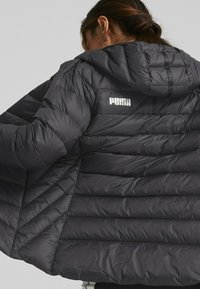 Sort pufferjakke med hætte, der har quiltet design, skinnende tekstur og et fremtrædende hvidt "PUMA" logo på ryggen.