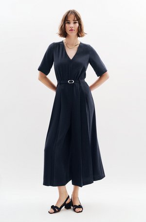 Kobieta ubrana w czarny kombinezon z paskiem, szerokimi nogawkami i rękawami do łokci, w połączeniu z czarnymi sandałami na obcasie i złotym naszyjnikiem z łańcuszka.