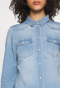 Helles blaues Denimhemd mit spitzem Kragen, Druckknöpfen und zwei Brusttaschen. Verfügt über eine glatte Textur und kontrastierende Nähte.
