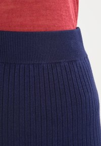 Detailní záběr pasu oblečeného v červeném topu zasunutém do vysoce pasové, pruhované pletené sukně tmavě modré barvy.