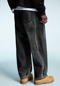Volné černé džíny z denimu s viditelným prošíváním, dvěma zadními kapsami a mírně vybledlou strukturou. Spárováno s hnědými pracovním botami.