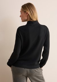 Schwarzes Oberteil mit Stehkragen und langen Ärmeln, aus glattem Stoff gefertigt. Der Rücken hat einen leichtem Kragen und ein tailliertes Design. Getragen mit grauen Hosen.