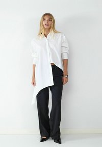 Weißes Hemd mit asymmetrischem Saum, kombiniert mit schwarzen, weit geschnittenen Hosen und schwarzen Schuhen mit spitzer Zehe. Insgesamt minimalistisches Design.