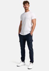 Jeune homme debout les mains dans les poches, portant un t-shirt blanc, un pantalon cargo bleu marine et des baskets blanches sur un fond uni.