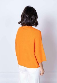 Maglione arancione a costine con maniche ampie, visto da dietro. Indossato con pantaloni bianchi, caratterizzati da una vestibilità svasata e un tessuto testurizzato.