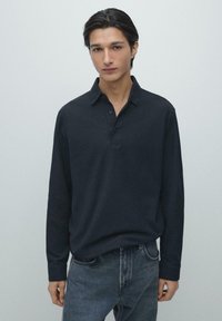 Marineblaues Langarm-Poloshirt mit Kragen, Knopfleiste und strukturierter Oberfläche, kombiniert mit dunklen Jeans. Modell wird von der Taille aufwärts gezeigt.