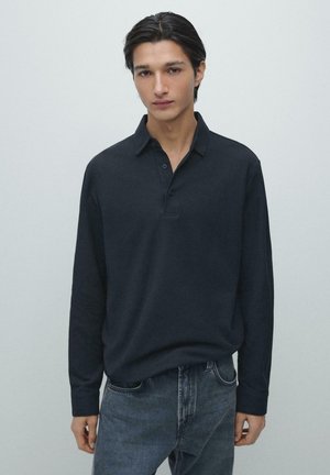Marineblaues Langarm-Poloshirt mit Kragen, Knopfleiste und strukturierter Oberfläche, kombiniert mit dunklen Jeans. Modell wird von der Taille aufwärts gezeigt.